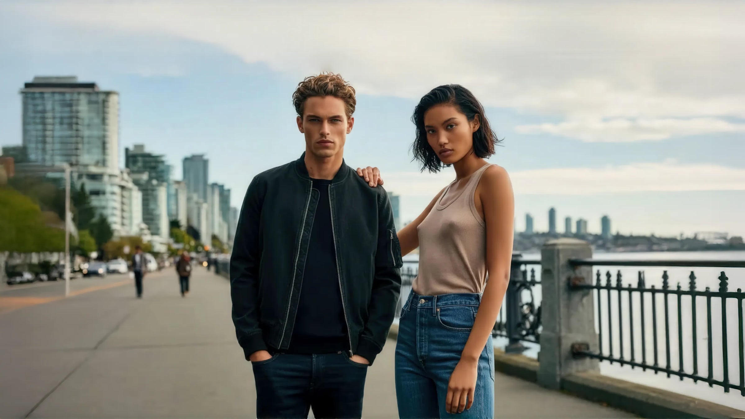 The image shows two individuals standing confidently on a wide pathway near a waterfront. The background features a modern urban skyline with tall buildings, suggesting a metropolitan setting. The male figure is wearing a black bomber jacket and dark jeans, while the female figure is dressed in a beige tank top and blue jeans. Both have serious, direct expressions and are positioned closely, with the female placing a hand on the male's shoulder.