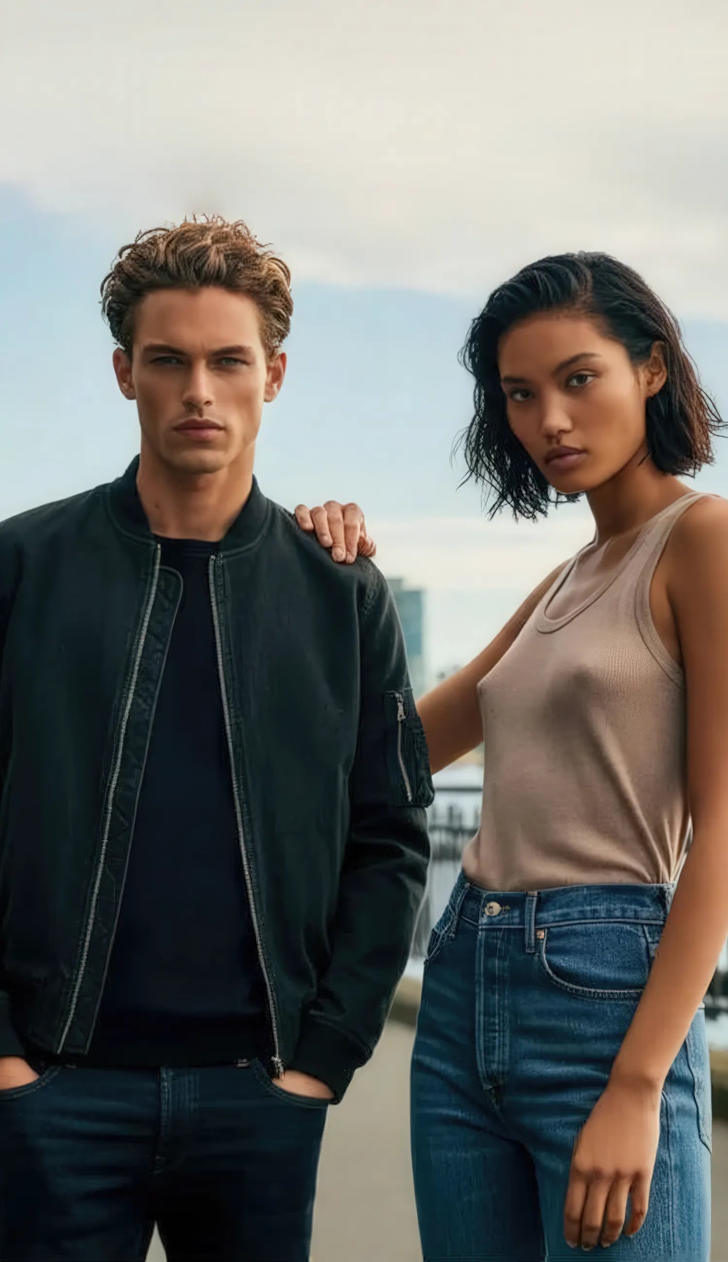 The image shows two individuals standing confidently on a wide pathway near a waterfront. The background features a modern urban skyline with tall buildings, suggesting a metropolitan setting. The male figure is wearing a black bomber jacket and dark jeans, while the female figure is dressed in a beige tank top and blue jeans. Both have serious, direct expressions and are positioned closely, with the female placing a hand on the male's shoulder.